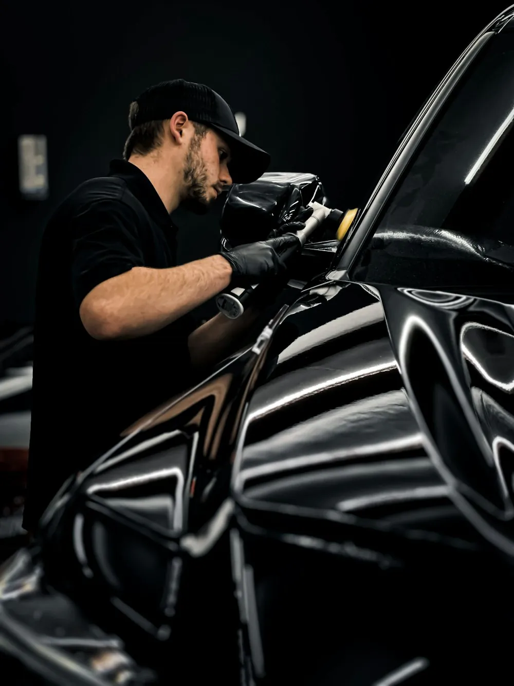 A detailer in black polishing a dark car body with a machine polisher