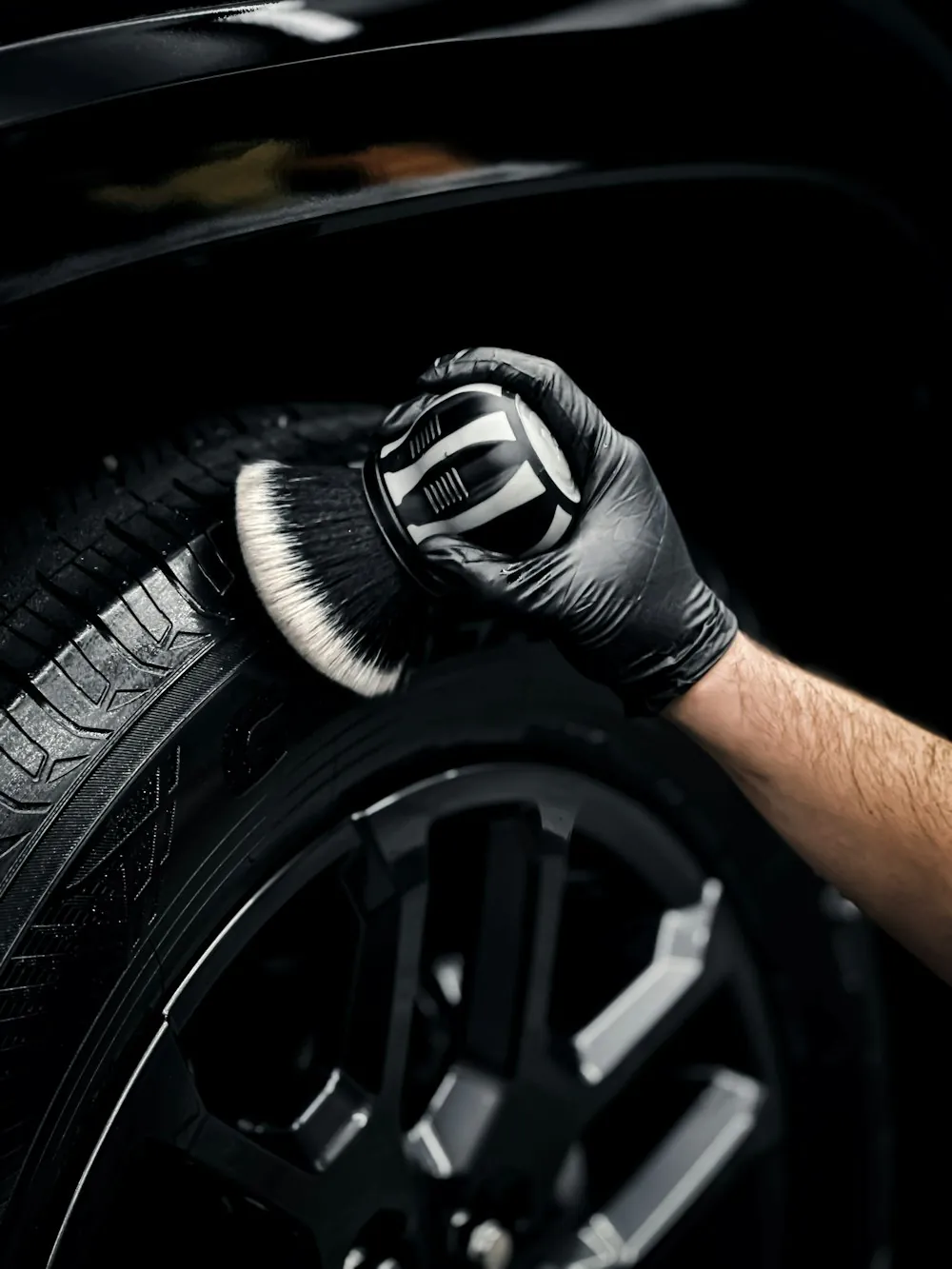 A gloved hand brushing the tire of a dark car with a detailing brush